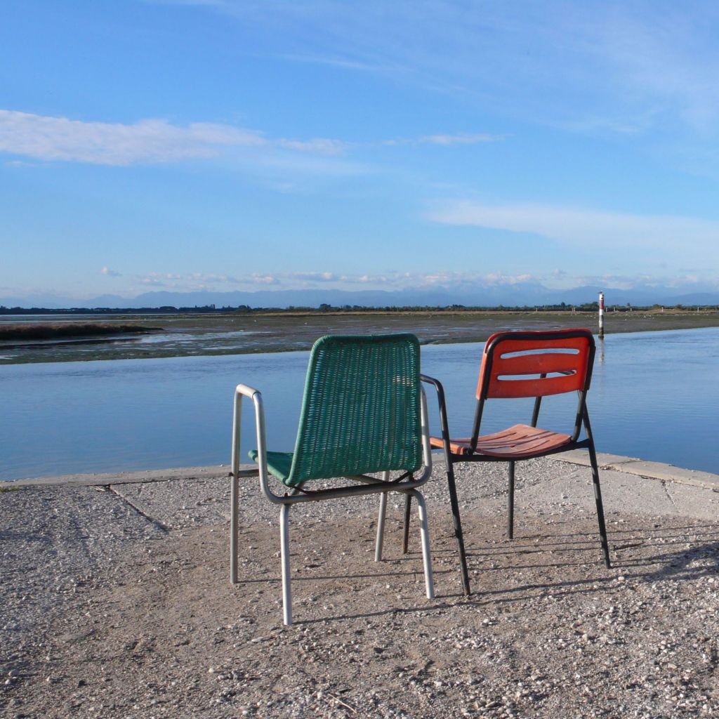 Ein grüner und ein oranger Stuhl stehen einsam an einer Kante und blicken über eine Wasserfläche in Richtung Berge am Horizont.